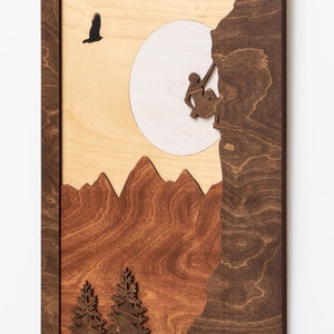 May include: Wooden wall art featuring a mountain climber, mountains, and trees. The artwork is framed in dark brown wood, with a light wood background. A silhouetted bird flies near a white circle, representing the sun.