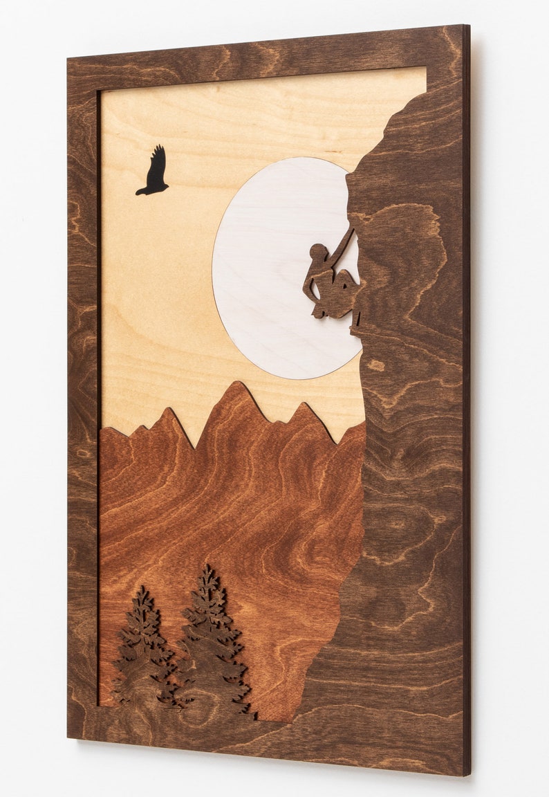 May include: Wooden wall art featuring a mountain climber, mountains, and trees. The artwork is framed in dark brown wood, with a light wood background. A silhouetted bird flies near a white circle, representing the sun.