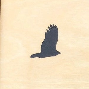 May include: A wooden plaque featuring a dark gray silhouette of a bird in flight. The bird is depicted with its wings spread, set against a light beige background. The plaque is framed with a dark brown border.