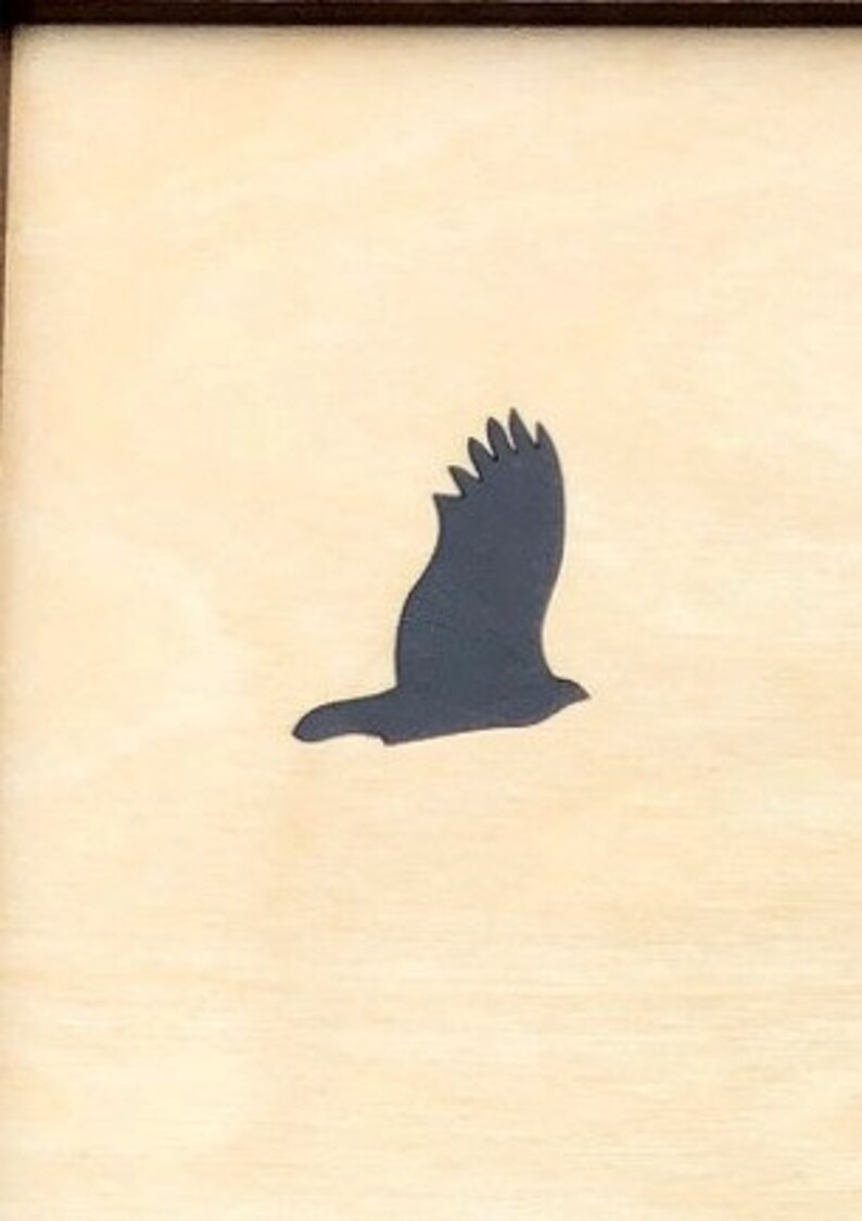 May include: A wooden plaque featuring a dark gray silhouette of a bird in flight. The bird is depicted with its wings spread, set against a light beige background. The plaque is framed with a dark brown border.