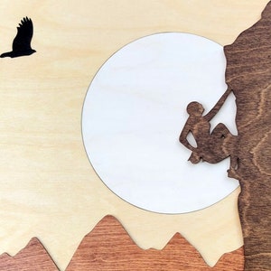 May include: Wooden wall art featuring a silhouette of a rock climber ascending a cliff face. A white circle represents the moon, with a silhouetted bird in flight. Brown mountains are at the bottom, all on a light wood background.