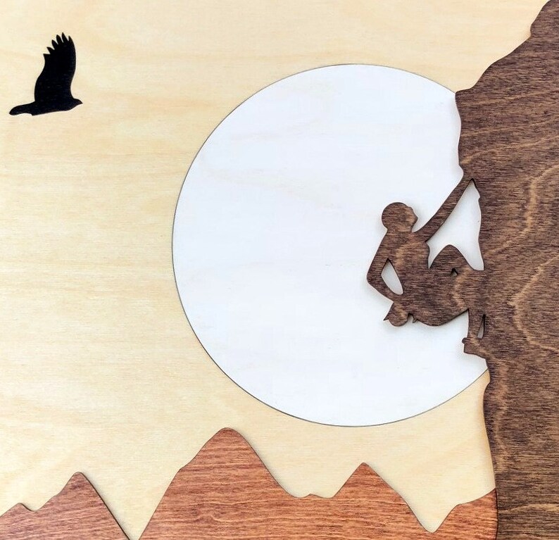 May include: Wooden wall art featuring a silhouette of a rock climber ascending a cliff face. A white circle represents the moon, with a silhouetted bird in flight. Brown mountains are at the bottom, all on a light wood background.