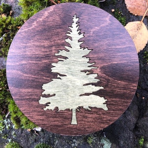 May include: A round wooden coaster with a laser-cut design of a pine tree in a light brown color. The coaster has a dark brown wood grain background.