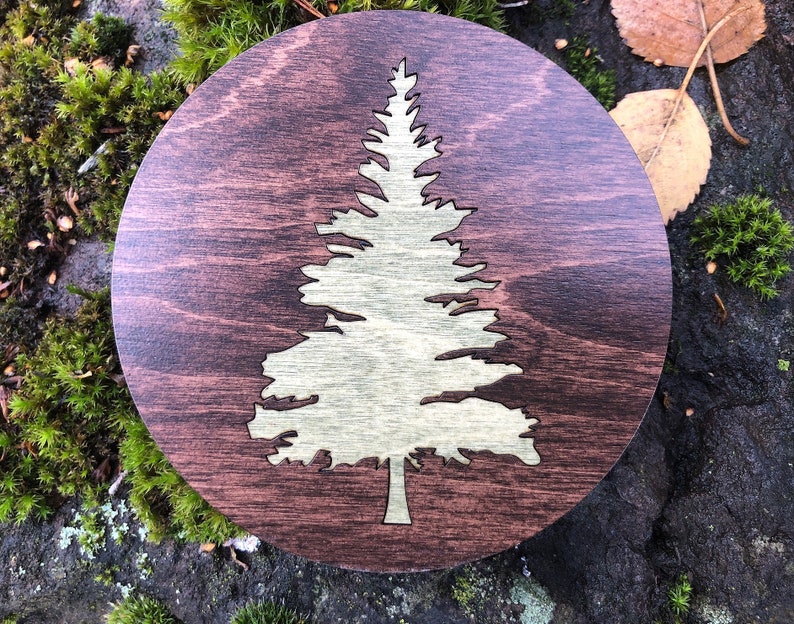 May include: A round wooden coaster with a laser-cut design of a pine tree in a light brown color. The coaster has a dark brown wood grain background.