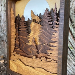 May include: A wooden wall art piece with a layered design featuring a wolf howling at the moon in a forest setting. The piece is framed in a dark brown wood frame.