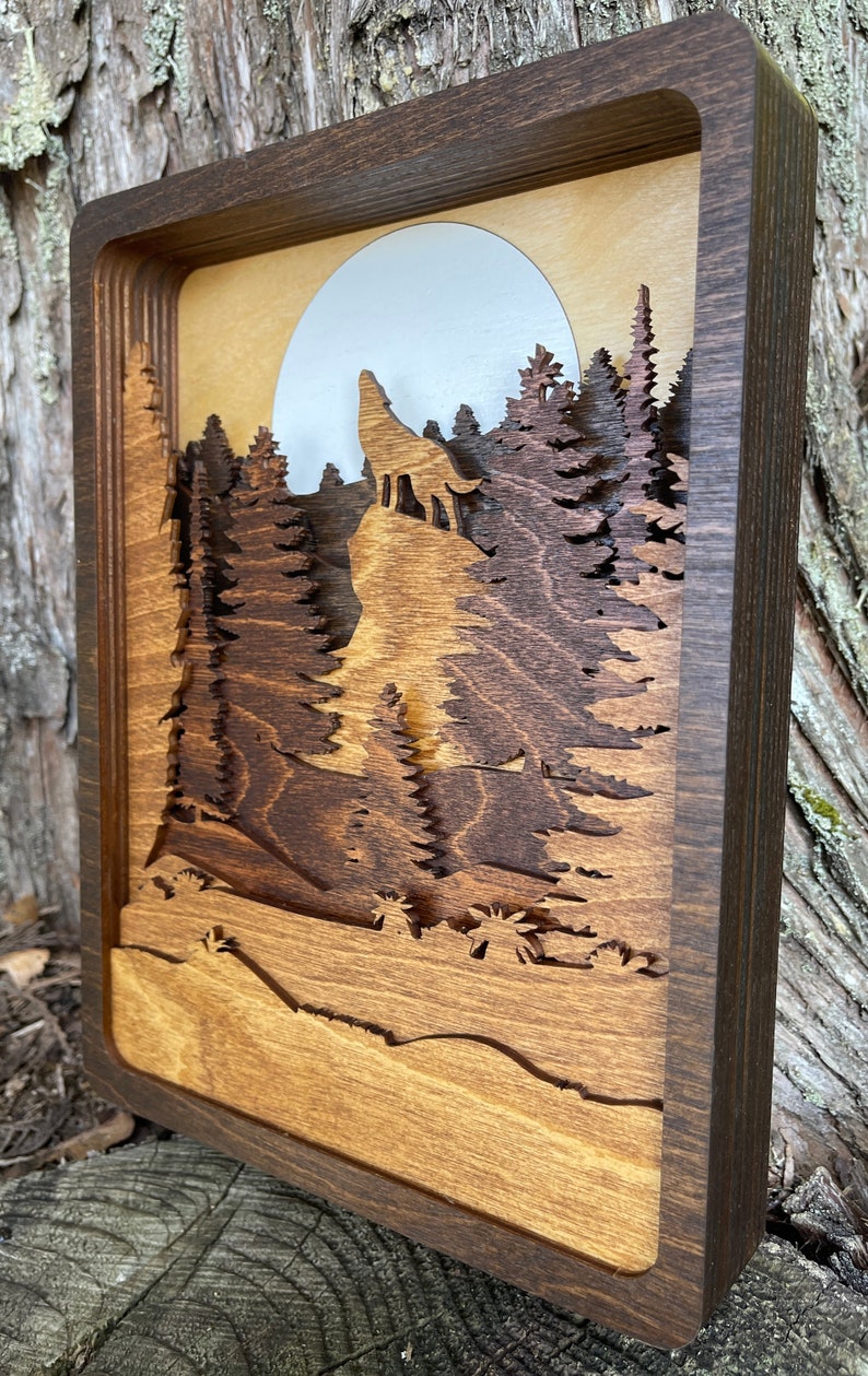 May include: A wooden wall art piece with a layered design featuring a wolf howling at the moon in a forest setting. The piece is framed in a dark brown wood frame.