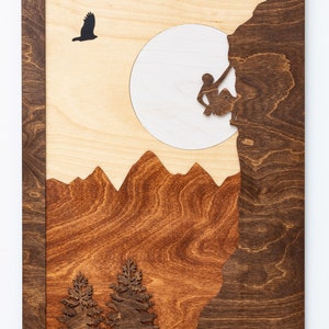 May include: Wooden wall art featuring a mountain climbing scene. The artwork depicts a climber, mountains, trees, and a soaring bird, all set against a light wood background with a dark wood frame. The design is a layered, laser-cut relief.
