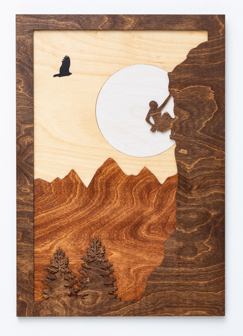 May include: Wooden wall art featuring a mountain climbing scene. The artwork depicts a climber, mountains, trees, and a soaring bird, all set against a light wood background with a dark wood frame. The design is a layered, laser-cut relief.