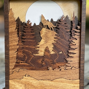 May include: A wooden wall art piece with a wolf howling at the moon in a forest setting. The piece is made of layered wood and has a dark brown stain.