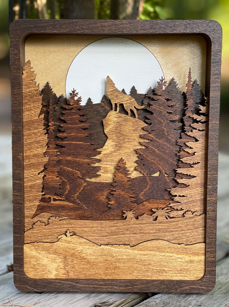 May include: A wooden wall art piece with a wolf howling at the moon in a forest setting. The piece is made of layered wood and has a dark brown stain.