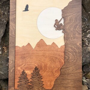 May include: Wooden wall art featuring a mountain climber on a rock face, a full moon, and a soaring bird. The artwork uses layered wood in shades of brown, tan, and orange, with a dark brown frame. The design includes mountain silhouettes and pine trees.