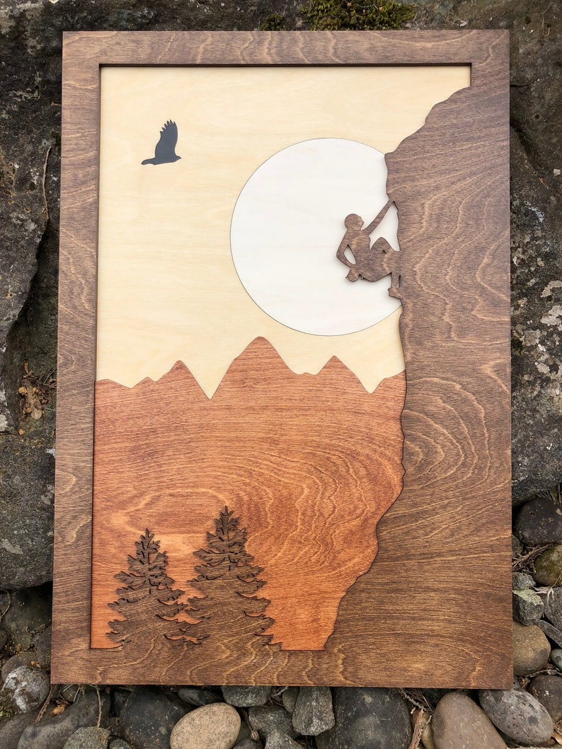 May include: Wooden wall art featuring a mountain climber on a rock face, a full moon, and a soaring bird. The artwork uses layered wood in shades of brown, tan, and orange, with a dark brown frame. The design includes mountain silhouettes and pine trees.