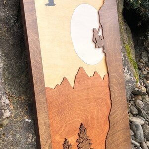 May include: Wooden wall art featuring a mountain climbing scene. The artwork depicts a climber, mountains, trees, a sun, and a flying bird. The piece is framed in a dark brown wood, with a layered design and a natural wood grain finish.