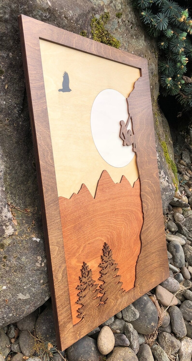 May include: Wooden wall art featuring a mountain climbing scene. The artwork depicts a climber, mountains, trees, a sun, and a flying bird. The piece is framed in a dark brown wood, with a layered design and a natural wood grain finish.