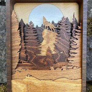 May include: A wooden wall art piece depicting a wolf howling at the moon in a forest setting. The wolf is silhouetted against a light blue moon, and the trees are carved in a layered style, creating a 3D effect.
