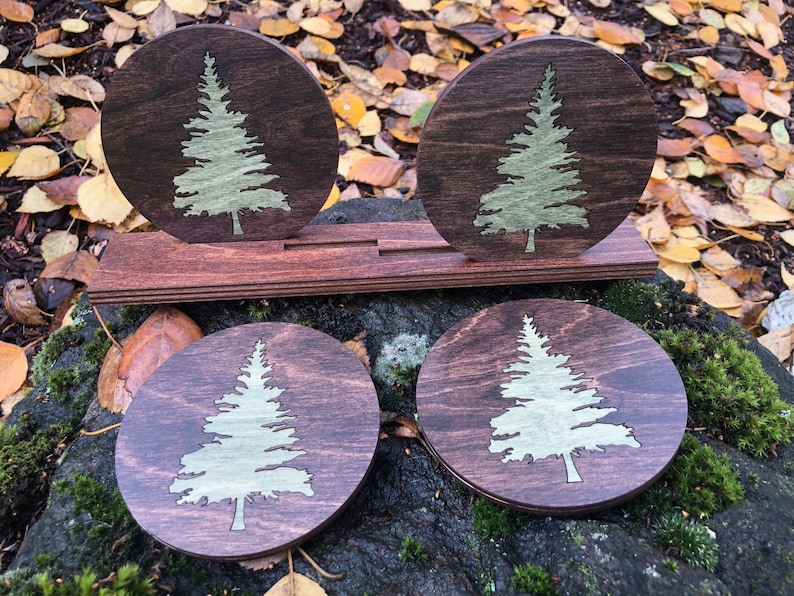May include: Four round wooden coasters with a laser-cut design of a green pine tree on a dark brown background. The coasters are arranged in a row on a wooden stand.
