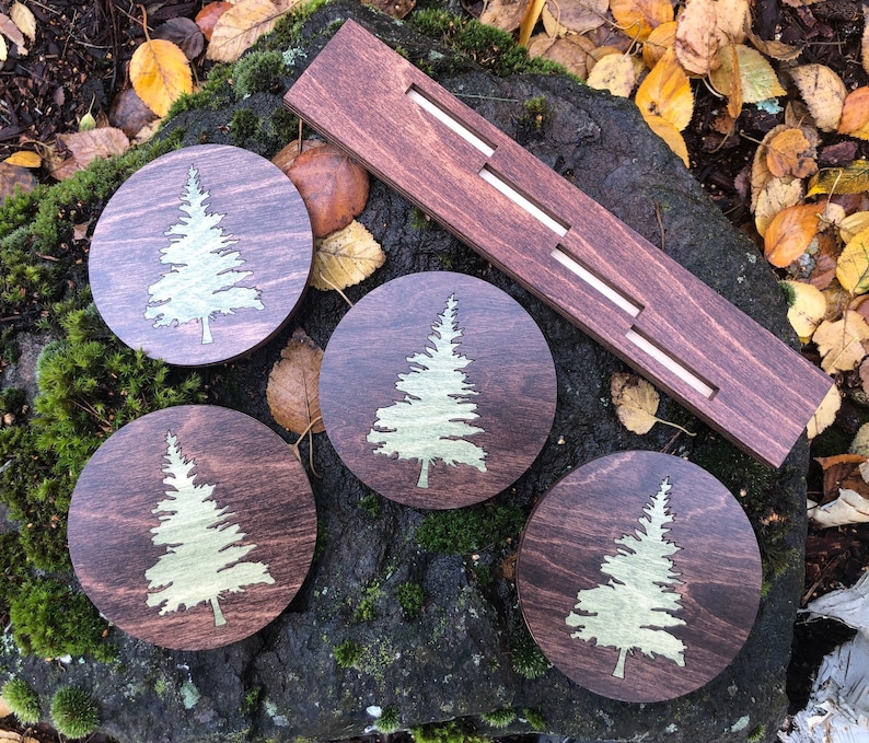 May include: Four round wooden coasters with a laser-etched design of a pine tree in gold on a dark brown background. The coasters are arranged in a circle around a wooden holder with three slots.