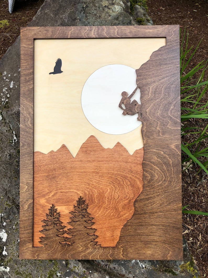 May include: Wooden wall art featuring a mountain climbing scene. The artwork has a layered design with a dark brown frame, a light brown background, and a silhouette of a climber, mountains, trees, a bird, and a full moon. The wood grain is visible.