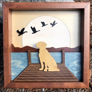 May include: A framed wooden artwork featuring a silhouette of a dog sitting on a jetty. The background depicts a large, white moon and several geese in flight.