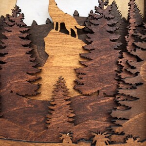 May include: A wooden wall art piece depicting a wolf howling at the moon in a forest setting. The wolf is silhouetted against a full moon, and the forest is made up of layered pine trees. The piece is made of dark brown and light brown wood.