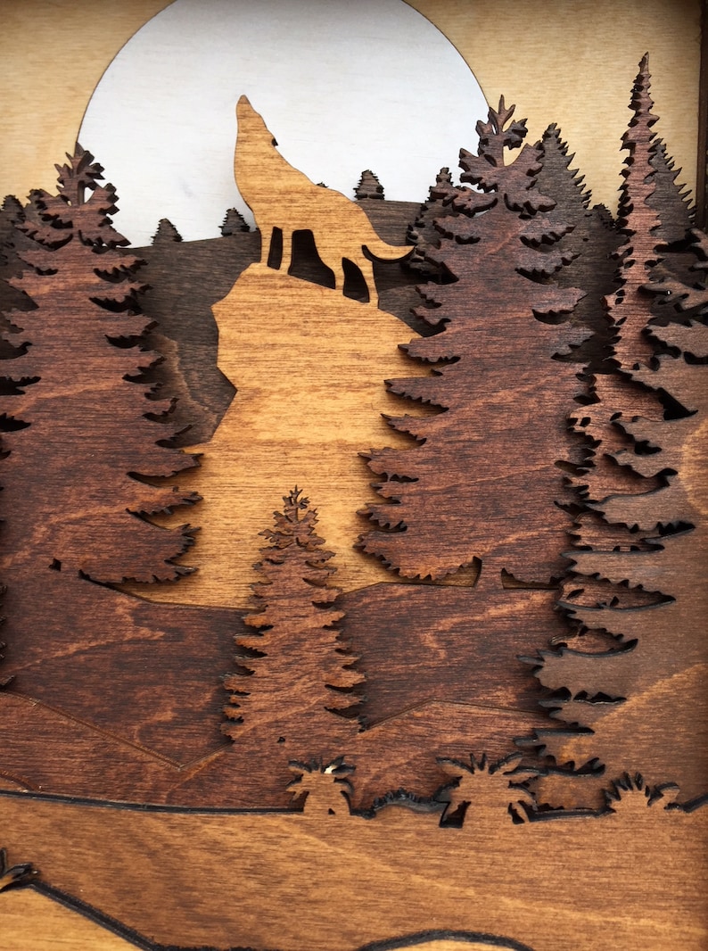 May include: A wooden wall art piece depicting a wolf howling at the moon in a forest setting. The wolf is silhouetted against a full moon, and the forest is made up of layered pine trees. The piece is made of dark brown and light brown wood.