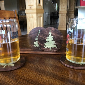 May include: Two glasses of beer on wooden coasters with a pine tree design. The coasters are dark brown with a light brown pine tree design.