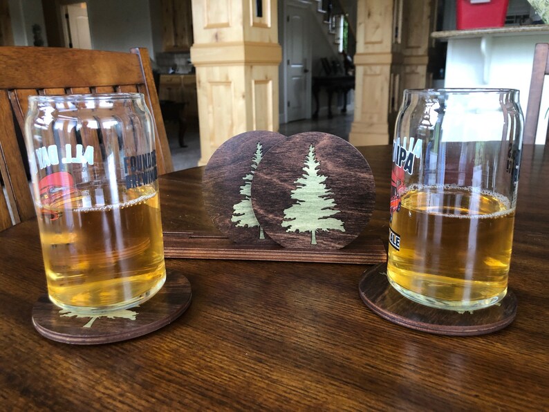 May include: Two glasses of beer on wooden coasters with a pine tree design. The coasters are dark brown with a light brown pine tree design.