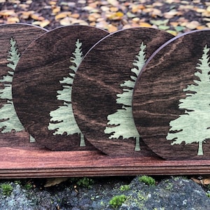 May include: Set of four round wooden coasters with a green pine tree design. The coasters are arranged in a row on a wooden stand.