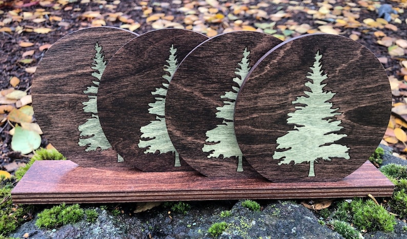 May include: Set of four round wooden coasters with a green pine tree design. The coasters are arranged in a row on a wooden stand.