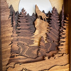 May include: A wooden wall art piece depicting a wolf howling at the moon in a forest setting. The wolf is silhouetted against a full moon, and the trees are carved in a layered style, creating a 3D effect.