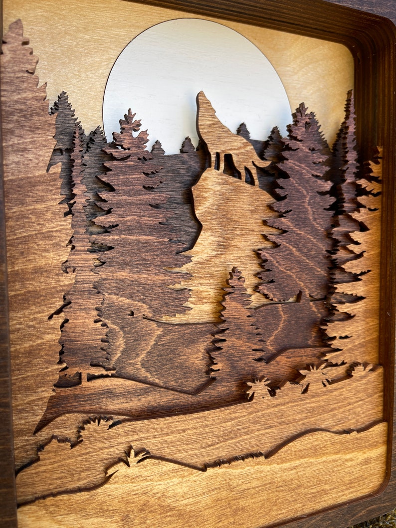 May include: A wooden wall art piece depicting a wolf howling at the moon in a forest setting. The wolf is silhouetted against a full moon, and the trees are carved in a layered style, creating a 3D effect.