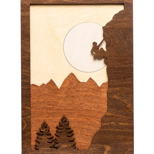 May include: Wooden wall art featuring a mountain climber silhouetted against a light-colored moon. The artwork has a dark brown frame and includes mountain and tree silhouettes in shades of brown and tan.