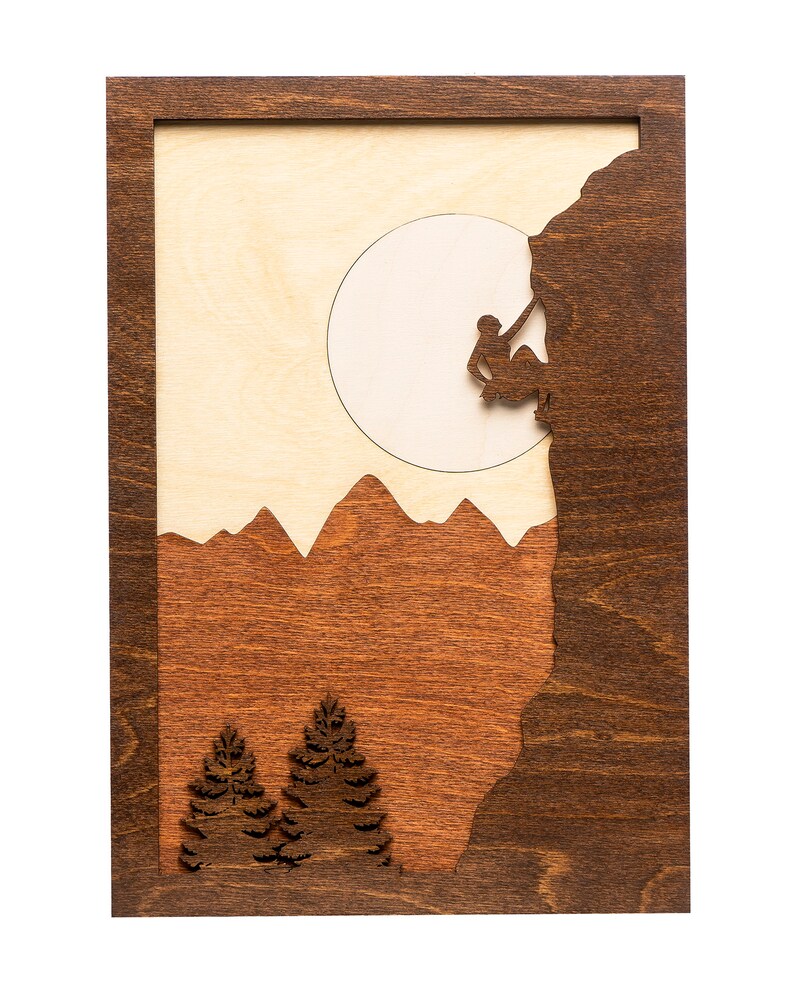 May include: Wooden wall art featuring a mountain climber silhouetted against a light-colored moon. The artwork has a dark brown frame and includes mountain and tree silhouettes in shades of brown and tan.