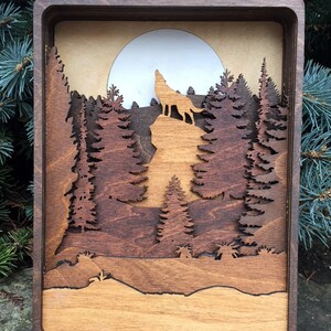 May include: A wooden wall art piece depicting a wolf howling at the moon in a forest setting. The wolf is silhouetted against a full moon, and the forest is made up of layered wood pieces to create a 3D effect.