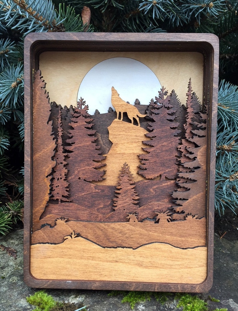 May include: A wooden wall art piece depicting a wolf howling at the moon in a forest setting. The wolf is silhouetted against a full moon, and the forest is made up of layered wood pieces to create a 3D effect.