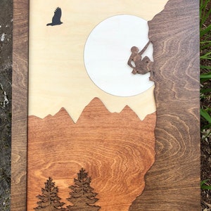May include: Wooden wall art featuring a mountain climbing scene. The artwork includes a climber, mountains, trees, and an eagle silhouette. The piece is framed in a dark wood, with a light wood background and a large white circle.