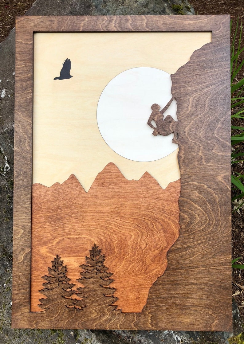 May include: Wooden wall art featuring a mountain climbing scene. The artwork includes a climber, mountains, trees, and an eagle silhouette. The piece is framed in a dark wood, with a light wood background and a large white circle.