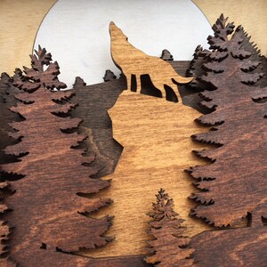 May include: A wooden wall art piece depicting a wolf howling at the moon in a forest setting. The wolf and trees are cut out of the wood, creating a layered effect.