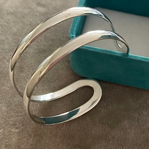 May include: A silver bangle bracelet with a double-band design. The bracelet is curved and has a smooth, polished finish.