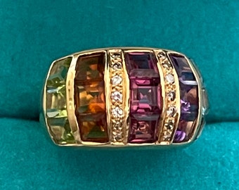 May include: A gold ring with a band of multi-colored gemstones. The ring features a row of green, orange, clear, purple, and blue gemstones.