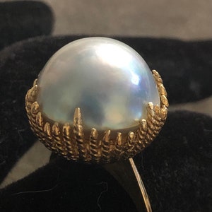 May include: A gold ring with a large, white pearl set in a gold claw setting. The ring is on a black background.