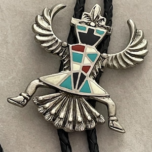 May include: Silver-toned bolo tie featuring a stylized Native American dancer with outstretched arms and legs. The dancer's costume is inlaid with turquoise, red, black, and white stones. The bolo tie has a black braided leather cord.