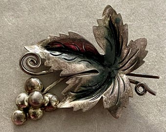 May include: A silver-toned brooch in the shape of a grape leaf and a cluster of grapes. The leaf has detailed veining and a curled tendril. The grapes are small and spherical. The brooch has a vintage aesthetic.