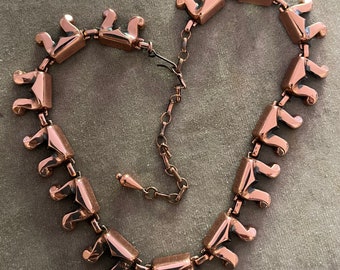 Puede incluir: Un collar de cobre con un diseño geométrico. El collar está hecho de piezas individuales de cobre que están unidas entre sí. El collar tiene un cierre.