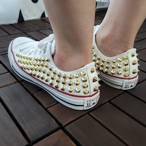 Puede incluir: Zapatillas blancas de lona con tachuelas doradas y el texto "Converse All Star" en el lateral.