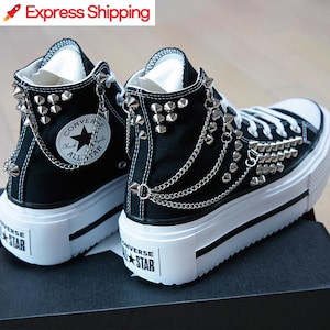 Puede incluir: Zapatillas altas negras con plataforma, tachuelas plateadas y detalles de cadena. Las zapatillas tienen suela blanca y el logo Converse All Star. El texto "Express Shipping" es visible en la esquina superior izquierda.