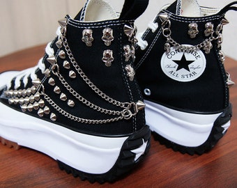 May include: Black high-top Converse sneakers with a platform sole. The shoes are decorated with silver studs, chains, and skull charms. The Converse All Star logo is visible on the side of the shoe.