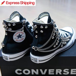 Puede incluir: Zapatillas Converse negras de caña alta con tachuelas y cadenas plateadas. Las zapatillas tienen una suela de goma blanca y una parte superior de lona negra. El logotipo de Converse es visible en el lateral de las zapatillas.