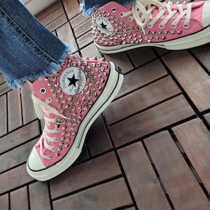 Puede incluir: Zapatillas Converse de caña alta de color rosa con tachuelas plateadas y un detalle de cadena. Las zapatillas están gastadas y tienen un aspecto vintage.