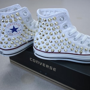 Puede incluir: Zapatillas Converse blancas de caña alta con tachuelas piramidales plateadas que cubren toda la zapatilla. El logotipo de Converse es visible en el lateral de la zapatilla.
